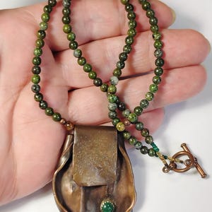 May include: A necklace featuring a strand of small, mottled green beads and a large, textured copper pendant. The pendant has a folded, organic shape with a single, small, round green stone set near the bottom. A copper toggle clasp secures the necklace.
