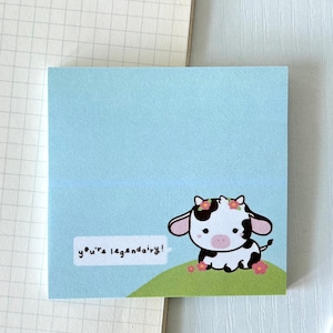 Puede incluir: Una nota adhesiva azul claro con una vaca de dibujos animados que lleva una corona de flores. La vaca está sentada en un parche de hierba verde con flores rosas. La nota dice "you're legendary!" en texto blanco.