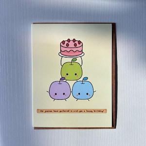 May include: A birthday card featuring a cartoon illustration. A pink cake sits atop a green apple, which is stacked on a purple and blue apple. The text reads: "the juninos have gathered to wish you a happy birthday!"
