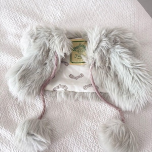 May include: A light gray faux fur shrug featuring a white satin lining with a repeating Cabbage Patch Kids logo print. The garment includes two long, thin, light pink velvet ties that hang down, each finished with a matching gray faux fur pom-pom.