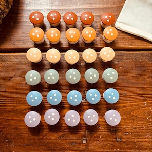 May include: A collection of twenty-five mushroom-shaped wooden knobs in various colors, including shades of orange, green, blue, and purple. Each knob features three white dots and a natural wood base. The knobs are arranged on a wooden surface.