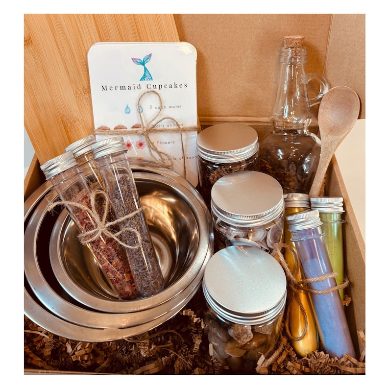 May include: A craft kit in a cardboard box containing two stainless steel bowls, a wooden spoon, a glass bottle, three jars with silver lids, and two glass test tubes filled with dried botanicals. A white card labeled Mermaid Cupcakes is included.