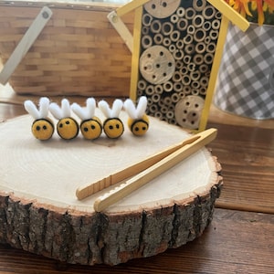 May include: Five yellow felt bumblebees with white wings are arranged in a row on a wooden surface. A pair of wooden tweezers are also on the surface.