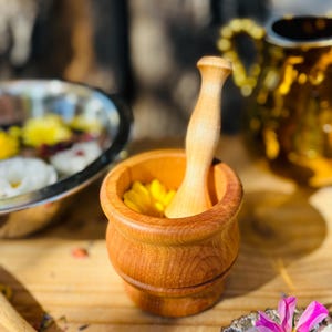 Pu&ograve; includere: Un mortaio e un pestello in legno con un fiore giallo all'interno del mortaio. Il mortaio e il pestello sono in legno marrone chiaro.