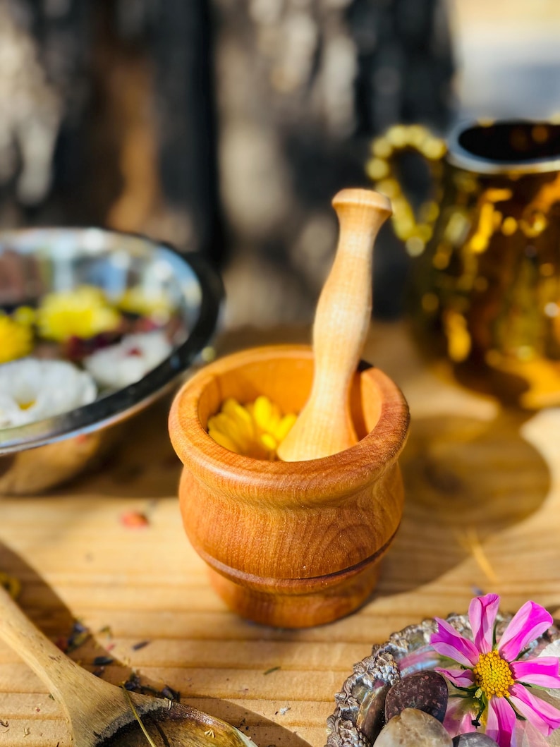 Pu&ograve; includere: Un mortaio e un pestello in legno con un fiore giallo all'interno del mortaio. Il mortaio e il pestello sono in legno marrone chiaro.