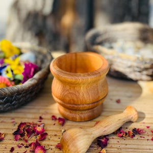Pu&ograve; includere: Un piccolo set di mortaio e pestello in legno. Il mortaio &egrave; di colore marrone chiaro e il pestello &egrave; di un marrone leggermente pi&ugrave; scuro. Il set &egrave; appoggiato su una superficie di legno con petali di rosa secchi sparsi intorno.