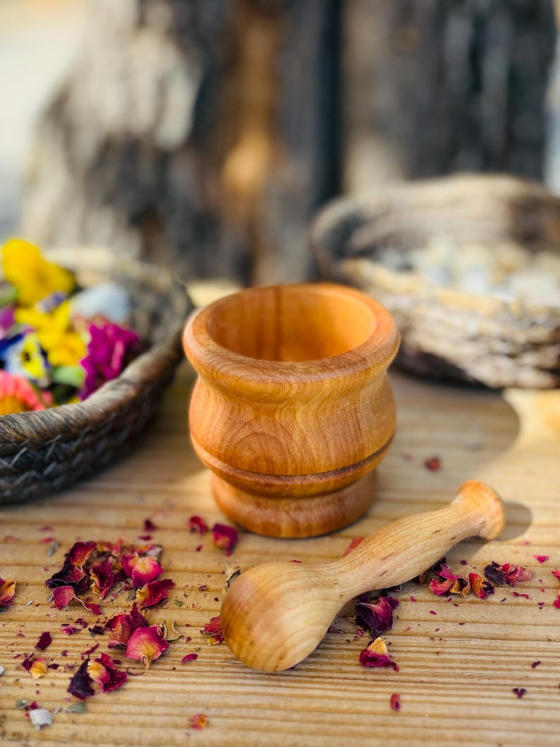 Pu&ograve; includere: Un piccolo set di mortaio e pestello in legno. Il mortaio &egrave; di colore marrone chiaro e il pestello &egrave; di un marrone leggermente pi&ugrave; scuro. Il set &egrave; appoggiato su una superficie di legno con petali di rosa secchi sparsi intorno.