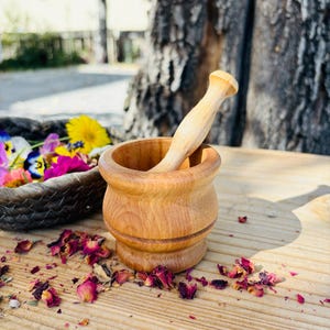 Pu&ograve; includere: Un mortaio e un pestello in legno su una superficie in legno con petali di rosa secchi sparsi intorno. Il mortaio e il pestello sono entrambi in legno marrone chiaro.