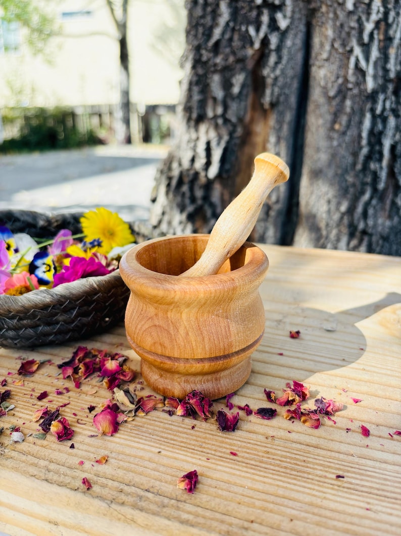 Pu&ograve; includere: Un mortaio e un pestello in legno su una superficie in legno con petali di rosa secchi sparsi intorno. Il mortaio e il pestello sono entrambi in legno marrone chiaro.