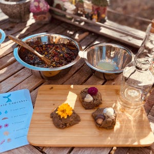 May include: A wooden cutting board holds three round, textured brown dough mounds topped with a yellow flower, a purple flower, and small seashells. Nearby are two stainless steel bowls, one containing a dark floral mixture with a wooden spoon.