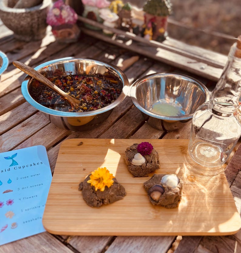 May include: A wooden cutting board holds three round, textured brown dough mounds topped with a yellow flower, a purple flower, and small seashells. Nearby are two stainless steel bowls, one containing a dark floral mixture with a wooden spoon.
