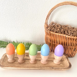 May include: Five pastel-colored wooden eggs in orange, yellow, green, blue, and purple, each in a wooden egg cup, arranged on a wooden tray. A wicker basket filled with brown paper and a fern are in the background.