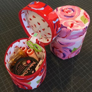 May include: Two cylindrical sewing kits. One is open, revealing sewing supplies like thread spools and scissors. The open kit has a red floral pattern on the outside and a strawberry print lining. The closed kit has a pink rose pattern.