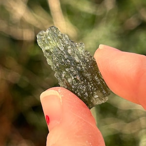 Pode incluir: Uma pequena pedra verde translúcida, de forma irregular, com uma superfície áspera e texturizada.