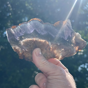 Pode incluir: Uma pedra preciosa natural bruta com uma aparência em camadas. A pedra apresenta tons de castanho, roxo e branco, com uma qualidade translúcida. As bordas são ásperas e irregulares, mostrando a forma orgânica da pedra.