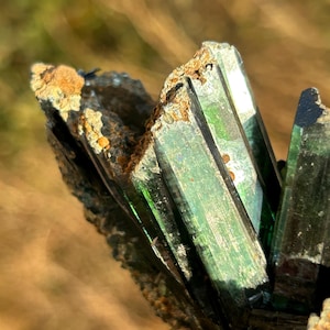 Pode incluir: Close-up de um aglomerado de cristais verde-escuros com um brilho metálico. Os cristais são afiados e angulares, com uma textura áspera e natural.