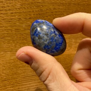 May include: A smooth, oval-shaped lapis lazuli stone with a deep blue color and white and gray swirls. The stone is polished and has a glossy finish.