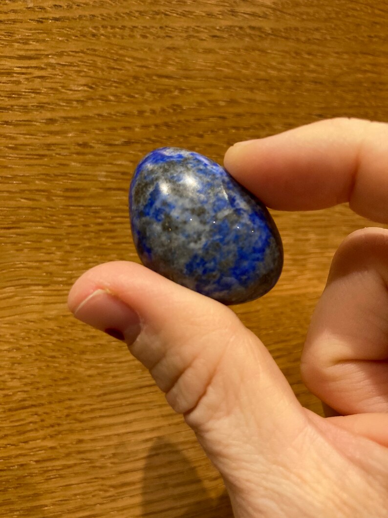 May include: A smooth, oval-shaped lapis lazuli stone with a deep blue color and white and gray swirls. The stone is polished and has a glossy finish.