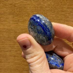 May include: Two smooth, oval-shaped lapis lazuli stones, one blue with white and gold flecks, the other a darker blue with white flecks, held in a hand.