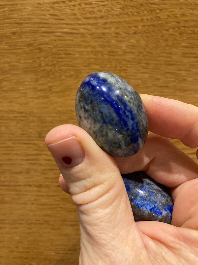 May include: Two smooth, oval-shaped lapis lazuli stones, one blue with white and gold flecks, the other a darker blue with white flecks, held in a hand.