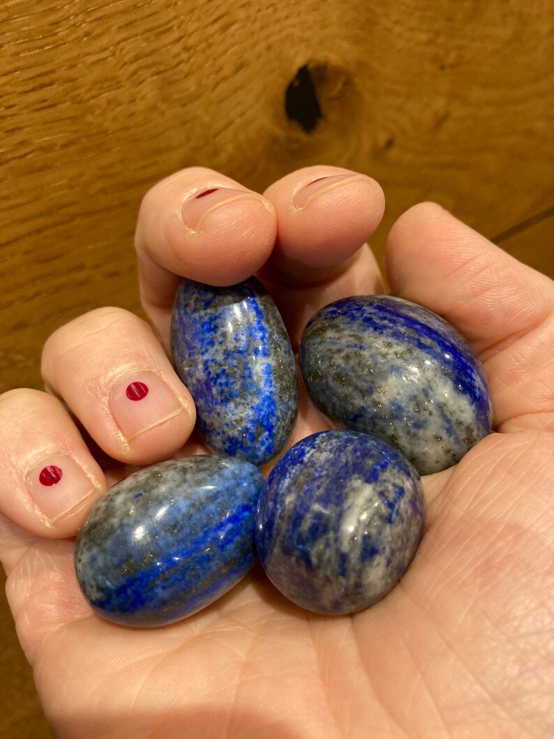 May include: Five smooth, oval-shaped lapis lazuli stones in various shades of blue with white and black marbling. The stones are held in a hand.