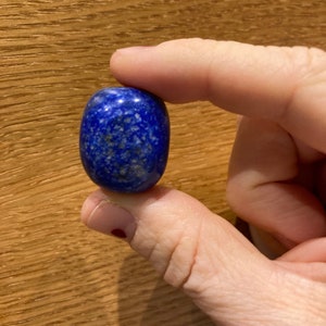 May include: A smooth, oval-shaped lapis lazuli stone with a deep blue color and white flecks. The stone is being held by a hand.