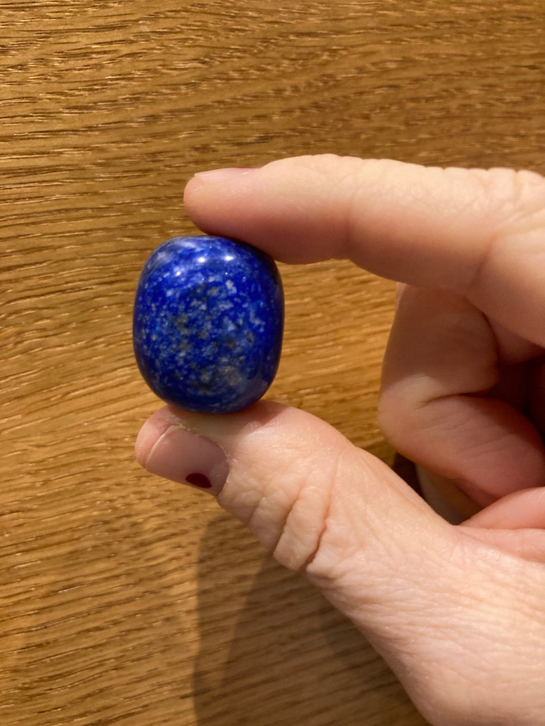 May include: A smooth, oval-shaped lapis lazuli stone with a deep blue color and white flecks. The stone is being held by a hand.