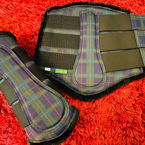 May include: A pair of horse leg boots featuring a dark blue, green, and orange plaid pattern. The boots have black interior lining, black hook-and-loop fastener straps, and a vertical strike plate. A small label on the larger boot reads FULL.