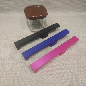 Pode incluir: Três puxadores de gaveta de cores diferentes. Os puxadores são de metal e têm forma retangular. As cores são preto, azul e rosa.