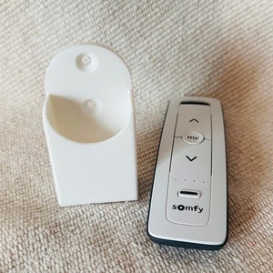 Może przedstawiać: Biały uchwyt ścienny i biały pilot z napisem "somfy" i "my". Pilot ma strzałki w górę i w dół oraz linię kropek.