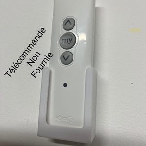 Peut inclure: Télécommande blanche avec trois boutons, dont deux avec des flèches et un avec le mot "my". La télécommande est dans un support mural blanc avec le mot "somfy" imprimé dessus.