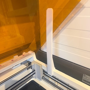 Op de afbeelding: Een witte 3D-printer steunarm bevestigd aan het interne frame van een machine met een doorschijnend amberkleurig deksel. De printer heeft een zwarte basisplaat, metalen geleiderails en een zwart distributieriemmechanisme aan de binnenkant.