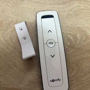 Op de afbeelding: Witte afstandsbediening met zwarte knoppen en de tekst "somfy" en "my". De afstandsbediening ligt naast een witte plastic wandbevestiging.