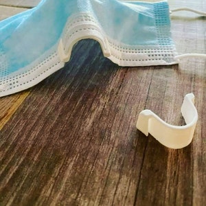 Op de afbeelding: Een witte plastic gezichtsmaskerhouder. De houder is U-vormig en is ontworpen om de bandjes van een gezichtsmasker vast te houden, zodat ze niet van de oren vallen.