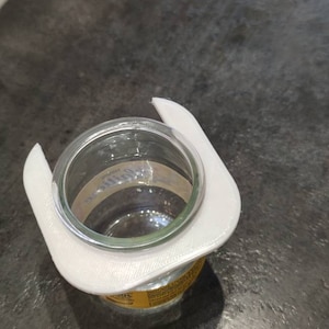 Könnte beinhalten: Weißer Plastik-Deckelhalter für Gläser. Der Halter ist so konzipiert, dass er um den Deckel eines Glases passt und ihn aufrecht hält.