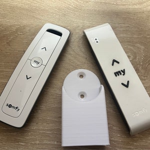 May include: Two white Somfy remote controls with black buttons and the word "my" on them. One remote control is attached to a white wall mount.