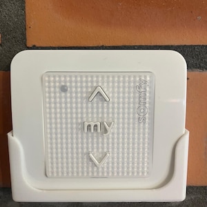 Może przedstawiać: Biały, montowany na ścianie pilot z białym panelem przycisków i czarnym przyciskiem. Panel ma czarny punkt, strzałkę skierowaną w górę, słowo "my", strzałkę skierowaną w dół i słowo "somfy".