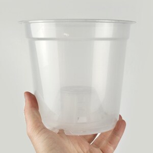 Peut inclure: Pot de plante en plastique transparent, avec un bord légèrement évasé. Le pot est tenu par une main sur fond blanc. Il a une forme cylindrique avec une base surélevée et des trous de drainage. Conçu pour les plantes.