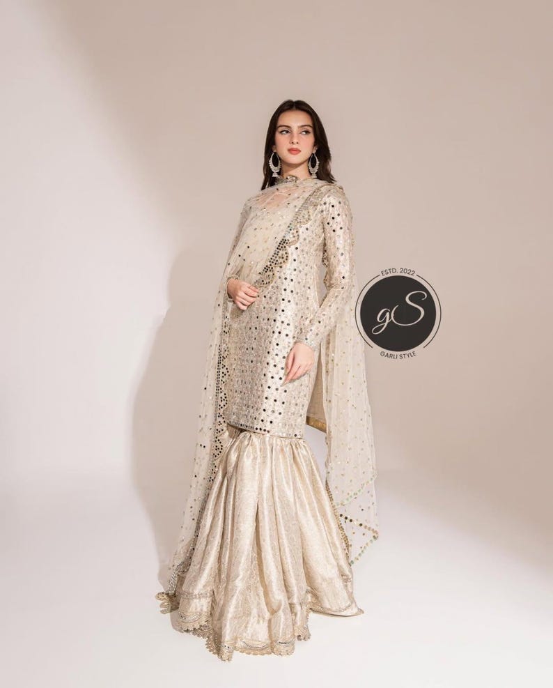 May include: A cream-colored, long-sleeved formal outfit with intricate embellishments. The ensemble includes a top with a high neckline and flared pants. A sheer, embellished shawl drapes over the shoulders. The outfit is adorned with small, reflective accents.
