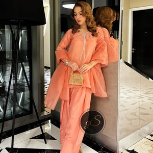 Peut inclure: Un salwar kameez de couleur p&ecirc;che avec un d&eacute;tail de n&oelig;ud blanc sur l'encolure. La tenue est associ&eacute;e &agrave; un dupatta assorti et &agrave; un petit sac &agrave; main de couleur or.