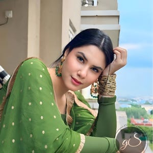 May include: A woman wearing a green polka dot dress with gold trim and gold bangles. She is looking at the camera and has her hair pulled back. The background is a blurry view of a city.