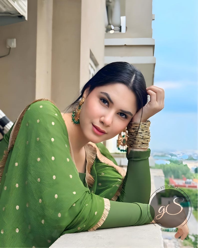 May include: A woman wearing a green polka dot dress with gold trim and gold bangles. She is looking at the camera and has her hair pulled back. The background is a blurry view of a city.