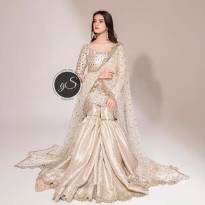 May include: A cream-colored, embellished formal outfit. The ensemble includes a long-sleeved top, wide-legged pants, and a sheer, dotted shawl. The outfit is adorned with intricate embroidery and sequins. The image includes a circular logo with the text "GS" and "ESTD. 2022".
