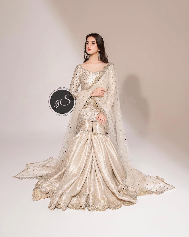 May include: A cream-colored, embellished formal outfit. The ensemble includes a long-sleeved top, wide-legged pants, and a sheer, dotted shawl. The outfit is adorned with intricate embroidery and sequins. The image includes a circular logo with the text "GS" and "ESTD. 2022".