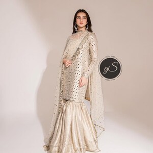 May include: A cream-colored, long-sleeved formal outfit with intricate embellishments. The ensemble includes a top with a high neckline and flared pants. A sheer, embellished shawl drapes over the shoulders. The outfit is adorned with small, reflective accents.