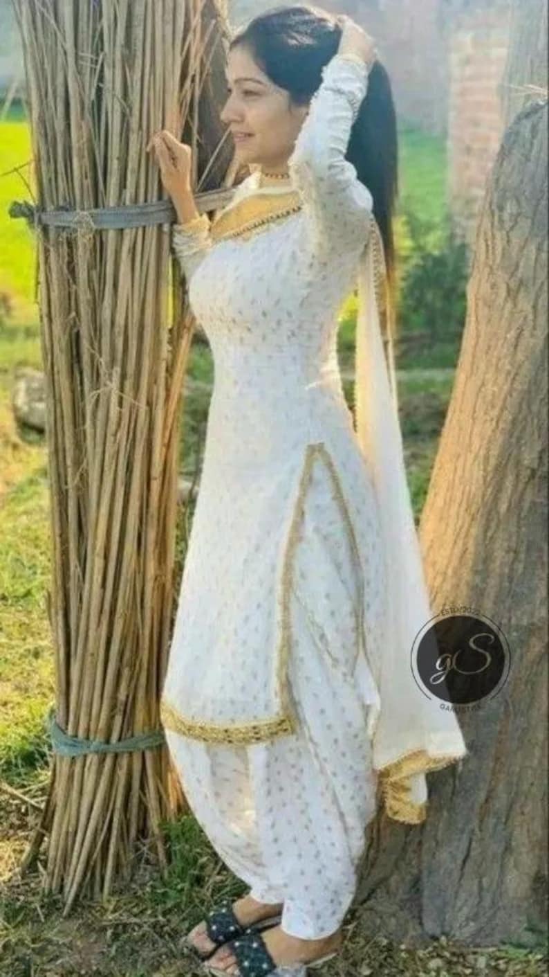 May include: A white salwar kameez with gold trim and a matching dupatta. The outfit features a subtle dotted pattern and is worn with black sandals. The background includes a bundle of sticks and a tree.