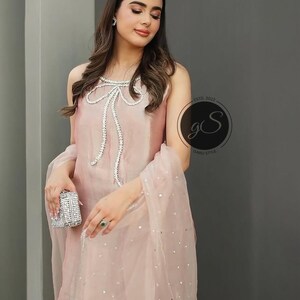 以下が含まれることがあります： 女性は、パールとラインストーンのボウディテールが付いたピンクのノースリーブトップを着ています。彼女はまた、銀色の装飾が施された透き通ったピンクのドゥパッタを着ています。トップとドゥパッタはどちらも柔らかく流れるような生地で作られています。
