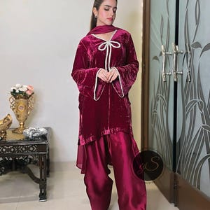May include: A burgundy velvet tunic with a white bow and pearl embellishments, paired with matching burgundy pants. The outfit has a flowing, elegant style.