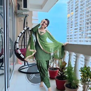May include: A woman wearing a green salwar kameez with gold trim and a matching green dupatta. She is standing on a balcony with a view of a city skyline.