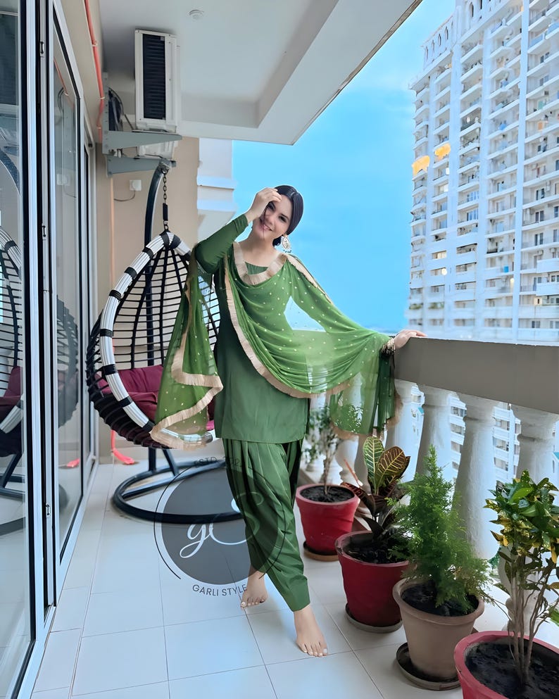 May include: A woman wearing a green salwar kameez with gold trim and a matching green dupatta. She is standing on a balcony with a view of a city skyline.
